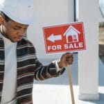 Młody fachowiec w kasku trzyma tablicę na sprzedaż przed budynkiem